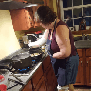 Quadruple amputee woman leaning over stove while cooking. She wears prosthetic legs and prosthetic brace on arm to cook with.