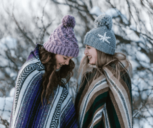 2 girlfriends wrapped in blankets, wearing hats and laughing, standing outside in winter